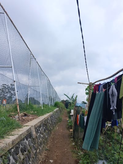 A narrow dirt path stretches into the distance between a tall chain-link fence on the left and a clothesline on the right. Laundry hangs from the line, partially obscuring a small shack. The fence runs atop a stone wall, with a grassy area and a soccer field visible behind it. Overcast sky dominates the scene. A lone power line descends diagonally across the upper-right portion of the view.