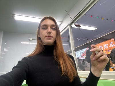 An indoor photograph captures a young adult female positioned centrally, looking directly at the camera. She has long, reddish-brown hair and is dressed in a black turtleneck sweater. Her right hand, featuring light pink fingernail polish, holds a long metal skewer with a dark, cooked food item impaled near its tip. A small white wrapper or tissue is also clutched in her right hand. The interior setting features white, possibly paneled or tiled, walls and ceiling with a fluorescent light fixture. A light green surface is visible in the bottom right. Through a large window behind the subject, an outdoor evening scene in Licheng District, China, is visible. This exterior view includes a string of multi-colored decorative lights (red, green, yellow, blue) and illuminated signs from street stalls or shops. A prominent orange sign displays the Chinese characters "奶南瓜" (Nǎi Nánguā - Milk Pumpkin) and the number "303". Blurred figures are discernible in the background, indicative of pedestrian activity.