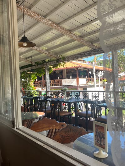 This image depicts a sunlit outdoor dining area and a view across to an adjacent building in the city of Cairu, Brazil. The scene is observed from an interior position, framed by a partially open white window on the left and a translucent white curtain on the right.

In the immediate foreground, a white table-tent display, featuring an image of three cocktail glasses, sits on a glass-topped table. A carved wooden chair back is partially visible.

The central focus is an empty outdoor dining terrace, covered by a white wooden ceiling with exposed beams, some exhibiting distressed paint. Two black industrial-style pendant lights with exposed bulbs hang from the ceiling. Green foliage, possibly vines, grows along the ceiling beams. The terrace is furnished with multiple unoccupied tables, each with a dark frame and a clear glass top, accompanied by matching dark chairs with light brown cushioned seats. Small vases containing dried plant material are present on some tables. A black wrought-iron railing with decorative scrollwork defines the edge of this dining area.

Beyond the terrace, across what appears to be a narrow street or walkway, stands another commercial building. This building features a tiled roof, exposed wooden support structures, and a balcony. A prominent sign on its facade reads "MERINOS" with "Restaurante | Pizzaria | Bar" underneath. A white pole stands between the two establishments. The background shows additional buildings, dense green trees, and a clear blue sky, indicating bright daylight conditions. No people are visible in the scene.