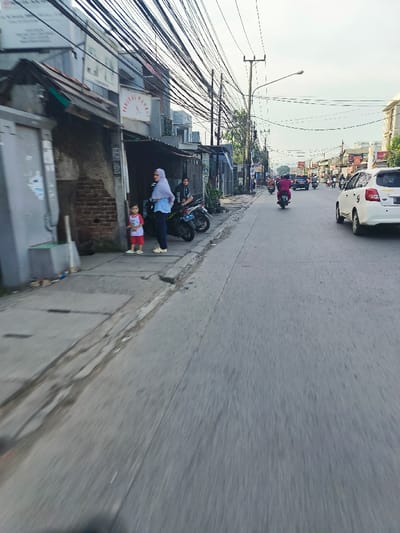 The image shows a street scene in Bojongsoang, Indonesia, located at latitude -6.978 and longitude 107.635. The street is paved with gray asphalt and has a sidewalk on the left side. There are several buildings along the street, including a two-story building with a sign that reads "WARUNG JAMU." The sidewalk is lined with a few trees and shrubs. There are a few people walking on the sidewalk and several motorcycles parked along the street. One motorcycle is being driven down the street by a person in a pink shirt. A white car with a black stripe along the side is parked on the street. The sky is a light blue color with some clouds. There are numerous power lines strung along the street. There are two adults and one child standing on the sidewalk. The adults are wearing a headscarf and a blue shirt, and the child is wearing a red shirt. The adults and child are standing in front of the store. There are several motorcycles parked along the street. The scene appears to be a typical street scene in an urban area. The image was likely taken by a person who was driving down the street.