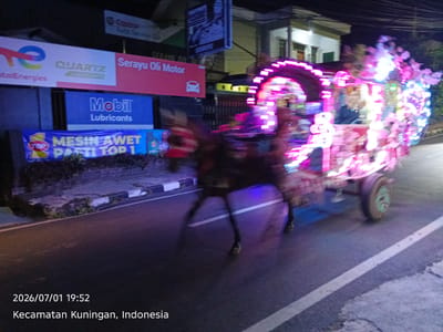 This is a nighttime photograph taken in Kuningan, Indonesia. The main subject is a horse-drawn carriage, or "delman," that is heavily decorated with colorful, blinking lights. The carriage appears to be moving, as evidenced by the motion blur of the horse and the carriage itself. The horse is a dark color, and its legs are visible as it walks on the asphalt road.
In the background, there are buildings and various signs. One prominent sign displays "Mobil Lubricants" in white text on a blue background. Another sign is red and reads "Serayu Oli Motor." There's also a sign with a large "e" logo and "TotalEnergies" written on it. A smaller sign above that mentions "Castrol Auto Service." The overall scene suggests a festive or celebratory event taking place after dark. The time stamp at the bottom indicates the photo was taken on July 1, 2026, at 7:52 PM. The road has white lines, and there's some vegetation and concrete barriers along the side of the road. The atmosphere is illuminated by the bright lights of the carriage and the surrounding signs.