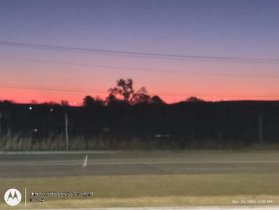 The image depicts a vibrant sunset scene. The sky transitions from a deep purple at the top to a fiery orange near the horizon. A dark, silhouetted treeline and some shrubbery span the middle ground. In the foreground, a gray road and grassy verge create a simple composition. The text indicates the photo was taken on a Motorola stylus 5G in 2024 and labeled "Dock-si" on November 26, 2025, at 6:20 AM.