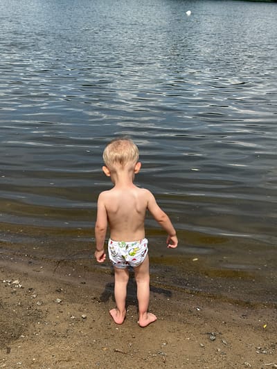 A young boy with blond hair stands on a sandy shore next to a body of water. He is wearing white swim trunks with a pattern of cartoon characters. The boy is facing away from the camera, looking out at the water. The sun appears to be shining, creating reflections on the water's surface. The shore is made up of sand and small pebbles. The water is calm with gentle ripples. The image appears to have been taken on a clear day, likely during the daytime. There is no visible text in the image. The setting is suggestive of a beach or lakeside environment.