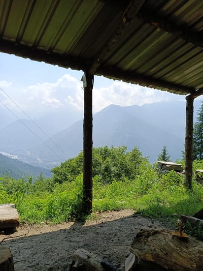 The image captures a view from under a corrugated metal roof supported by rough-hewn logs, overlooking a lush green mountainside and a hazy valley with a town nestled within it. The foreground shows a dirt path with scattered logs and a wooden bench on the left. A rustic wooden structure, possibly a shelter or resting spot, is the primary focus. In the middle ground, dense green vegetation covers the slope, punctuated by a few scattered trees. Beyond the immediate vegetation, the landscape transitions to rolling mountains that fade into a soft blue haze due to atmospheric perspective. A town or village is visible in the valley, with buildings and roads discernible, though softened by distance and haze. Power lines stretch diagonally across the scene. The sky is bright with scattered white clouds, suggesting a clear day. The overall impression is one of a serene, natural setting, likely in a rural or mountainous region of India, possibly near Sarsai, given the description. There are no people or animals present in the image.