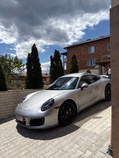 The image depicts a silver Porsche 911 parked in a driveway in what appears to be the outskirts of Dolno Drianovo, Bulgaria. A partial view of a brick building is visible across the driveway. There is at least one individual standing near the front of the car. The sky is partly cloudy. The driveway is paved with light-colored rectangular pavers. Tall, thin trees are present in the landscape. A license plate is visible at the front of the car, and it indicates the car is registered in Bulgaria.
