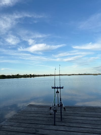 The image depicts two fishing rods placed in rod holders on a wooden dock. The scene is located in Khryashchevka, Russia. A body of water extends toward the horizon, with the opposite shoreline visible in the distance. The sky is light blue with wispy white clouds. There are no people present in the image.