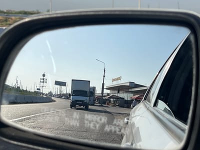 A reflection is visible in a car's side mirror, showing a road and a gas station. The reflection shows a bright, clear sky. The road is paved and appears to be a highway with multiple lanes. There are several vehicles on the road, including a white van and a truck. In the background, there is a gas station with a sign that reads "SHOX PATIR". There are also some buildings and infrastructure visible, such as power lines and streetlights. The scene appears to be in a suburban or rural area.