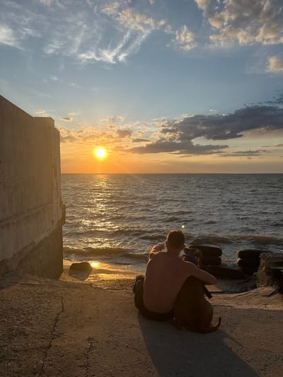 A man and his dog are sitting on a concrete embankment overlooking the sea at sunset. The man is shirtless, with his arm around the dog. The dog is a medium-sized, brown breed. The sun is setting over the water, casting a golden glow on the scene. There is a large concrete structure on the left side of the image, and in the background, there are some tire-like objects and debris. The sea is calm with small waves. The sky is blue with scattered clouds, illuminated by the setting sun. This scene is located in Zatoka, Ukraine.