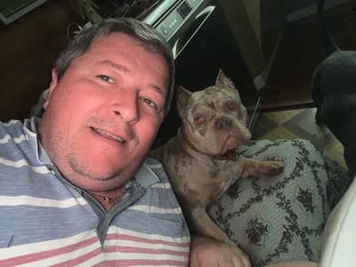 This is a close-up, overhead selfie taken in what appears to be a dimly lit room, possibly a living room or bedroom. The camera is held low, capturing the subjects from an unusual angle.

The primary subjects are a man and a dog, positioned close together. The man, with short graying hair and stubble, is smiling at the camera, his face tilted. He is wearing a blue and white striped shirt with red accents and a gold chain. Next to him, lying down, is a dog with distinctive pale fur and red eyes, looking towards the camera. The dog is wearing a brown collar. They appear to be relaxing together.

The background includes a patterned fabric, possibly a blanket or cushion, with a floral design in muted colors. Parts of what looks like a piece of furniture or an appliance are visible at the bottom of the frame, with some buttons and a screen suggesting electronic equipment. The floor tiling is also partially visible.

The overall mood is casual and intimate, suggesting a relaxed moment between a pet owner and his dog. The lighting appears to be artificial, indicating it could be evening or nighttime. There is no visible text in the image. The location context provided is Zelenogradskiy, Russia.