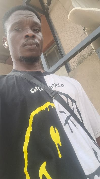 One male individual is depicted in a close-up, upward-angled shot in Badiko, Nigeria. The subject has dark skin, short dark hair, and minimal facial hair, appearing to be in his twenties or thirties. He is wearing a white wireless earbud in his left ear and looks directly into the camera with a neutral expression. He wears a short-sleeved t-shirt that is split down the middle: the left side is black with yellow dripping designs resembling a smiley face and text that reads "SMILE WORLD"; the right side is white with black dripping designs. A black shoulder bag strap, partially visible with "NICQUE" text, crosses diagonally over his chest. The background features a textured, light brown building wall. A dark metal railing or structure is visible horizontally across the mid-ground, with a window behind the subject's left side and a white corrugated surface visible in the upper right, consistent with an outdoor urban or residential environment.