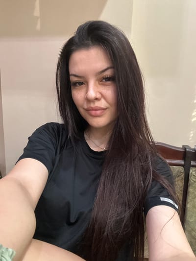 The image shows a young woman with long, dark brown hair and fair skin with freckles. She is wearing a black t-shirt and is looking towards the camera. The background appears to be an indoor setting with a beige-colored wall. A portion of a wooden chair with green upholstery is visible on the right side of the image. The lighting suggests it is daytime, possibly indoors with natural light from a window. The image quality is typical of a casual selfie. The specific location in the city of Sliven, Bulgaria, cannot be determined from the visual information provided.