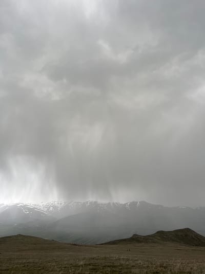 This landscape photograph shows a cloudy sky taking up much of the left side and some of the top of the image. On the right is a mountain range and on the far right is a grassy hillside. The location is identified as Hartagyugh, Armenia.