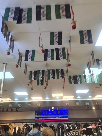 The image depicts the interior of a commercial establishment, likely a clothing store, located in Al-Qadam Municipality, Syria. The overhead white ceiling is extensively decorated with numerous small rectangular flags, each featuring three horizontal stripes (black, white, green from top to bottom) with three red stars on the white stripe, consistent with the flag of Syria. Interspersed among these flags are multiple artificial red roses suspended by strings. The ceiling is illuminated by an array of square and circular recessed light fixtures. Below the ceiling decorations, a blue neon sign displays the word "MONCLER" above another sign with horizontal red and white stripes. Below these signs, merchandise, primarily jackets and vests, is displayed on racks. Additional items, including shoes and hats, are visible on shelves to the right. Several individuals, observed from behind, are present in the lower portion of the frame, appearing to browse the displayed goods. One person wears a light brown head covering, another a blue denim jacket over a grey hooded garment, and a third a green hooded garment. No explicit interactions between individuals are discernible.