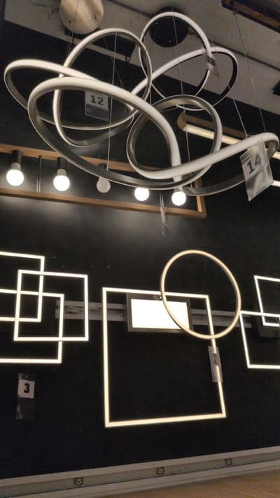 The image displays a retail environment, likely a lighting store, with several modern, illuminated fixtures. In the upper section, a large, multi-ringed pendant light with white LED elements is prominently featured, with price tags labeled "12" and "14" attached to it. Below this, a row of five exposed light bulbs hang from the ceiling, illuminating a wooden plank.
The lower section of the image showcases geometric LED lighting designs mounted on a dark wall. These include a series of interconnected squares and a circular ring light, all emitting a bright white glow. A price tag labeled "3" is visible near the bottom left, attached to a square fixture. Another tag with the number "2" is on the circular light. The overall impression is of a well-lit showroom displaying contemporary lighting solutions. There are no people visible in the image. The environment appears to be an indoor commercial space, with a hint of shelving or display units at the very bottom. There is no information in the image to suggest a specific location in the city of Sömmerda, Germany.