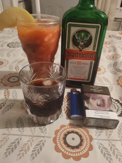 The image displays a table setting with alcoholic beverages and cigarettes. In the background, a green bottle of Jägermeister stands prominently, its label featuring a deer and the brand's name. In the foreground, to the left, a glass contains a dark liquid with ice. To the right of the Jägermeister bottle, a blue lighter is placed next to a pack of Sobranie Black cigarettes. A tall glass with a reddish beverage and a slice of lemon on the rim is visible behind the Jägermeister bottle, with a white straw inserted. The table is covered with a tablecloth featuring a floral and leaf pattern in orange, grey, and beige hues. The scene appears to be indoors, with soft lighting. The specific location in the city of Orbeli, Georgia, is not discernible from the visual information provided.