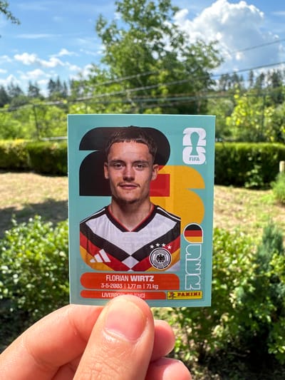 A hand is holding a soccer sticker featuring Florian Wirtz, a German player. The sticker is designed with a teal background and features Wirtz's portrait wearing a German national team jersey. The sticker also includes the FIFA logo, a large "2" and "0" graphic, and the Panini logo. The background of the image shows a lush green garden with trees and a blue sky with clouds, suggesting an outdoor setting. The location in Prisovo, Bulgaria is not explicitly visible or deducible from the provided image.