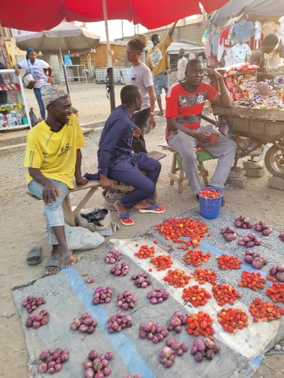 Four men are gathered near a vendor's wares at an outdoor market. Onions and what appear to be small tomatoes or peppers are displayed on a tarp on the ground. The men are casually dressed, some seated, while others stand nearby. One man wears a red shirt, another a yellow shirt, and another a blue outfit. A market cart with goods is visible in the background. Overall, it is a candid scene of social interaction amid the commerce of a marketplace.