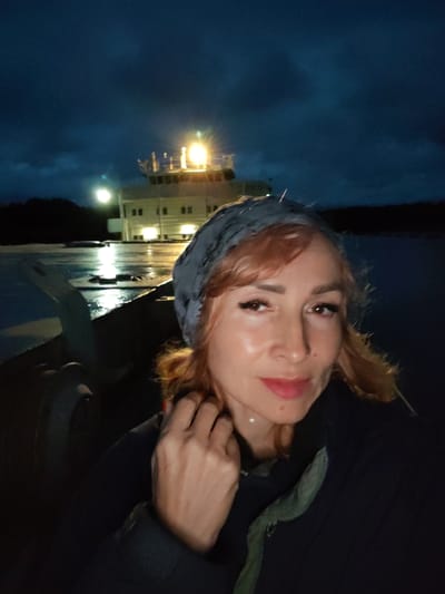 A woman is taking a selfie at night, possibly on a boat or dock. She has reddish-blonde hair, is wearing a grey headscarf and a dark jacket. Her right hand is touching her neck. Behind her, a building with lights on is visible, reflecting on a wet surface. The sky is dark blue and cloudy. The location is identified as Devyatiny, Russia.