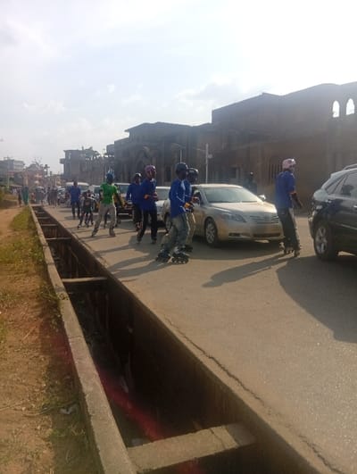 The scene depicts a daylight view of a street in Itakpe Junction, Nigeria. Approximately ten to fifteen individuals, primarily young males, are observed on rollerblades along an asphalt road. Many are attired in blue long-sleeved shirts, dark pants, and protective helmets (black, pink, or blue). One individual prominently features a bright green shirt, light-colored pants, and a green helmet with a distinct spiky crest. They are moving generally from left to right across the frame.

To the immediate left of the road, a concrete drainage ditch runs parallel, beyond which lies an unpaved dirt shoulder with sparse green vegetation. Several vehicles are present on the road alongside the rollerbladers, including a gold-colored sedan (possibly a Toyota Camry) positioned centrally and a dark-colored SUV partially visible on the far right. Other vehicles are visible further down the road in the distance.

In the background, on the right side of the road, stands a large, multi-story building with a weathered, dark facade and prominent arched windows, suggesting an older structure. Further left in the background, additional buildings, some appearing residential or under construction, are visible. The sky above is bright with some scattered clouds, and shadows on the ground indicate a clear day.