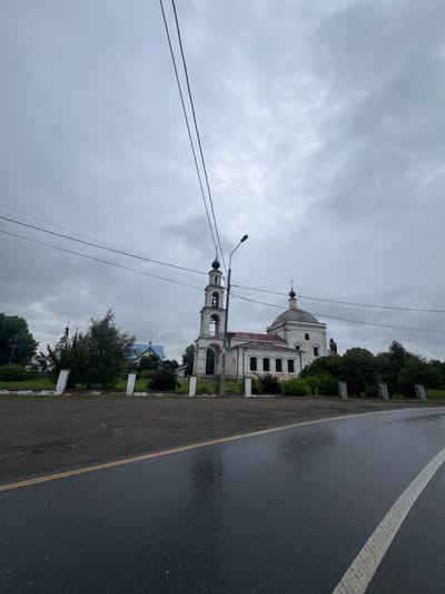 The image depicts a cloudy day in Десна Снт, Russia, with a church and its bell tower as the central focus. The road in the foreground is wet, suggesting recent rain. Electrical wires stretch across the sky, obscuring some of the view of the church and the surrounding trees. The church is made of white stone and has a dome with a cross on top. The bell tower is tall and slender, also made of white stone. A fence separates the road from the church grounds. There are no people or animals visible in the image. The overall atmosphere is somber due to the overcast sky.