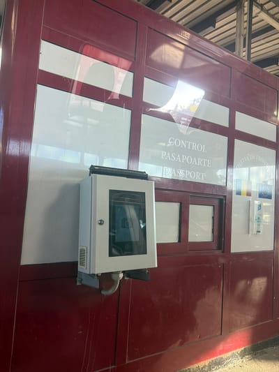 The image shows a border control booth in Sculeni, Romania. The booth is made of dark red metal and has several windows with white frosted glass. The words "CONTROL PASAPOARTE PASSPORT" are visible on one of the windows, indicating passport control. To the right of this text, the words "POLITIA DE FRONTIERA" are visible, along with the flags of Romania and the European Union. A white metal box with a screen is mounted on the left side of the booth. The screen displays an icon of a camera and the text "IDEMIA". The booth appears to be located outdoors, with sunlight reflecting on the surfaces. The ground in front of the booth is dusty and littered with debris.