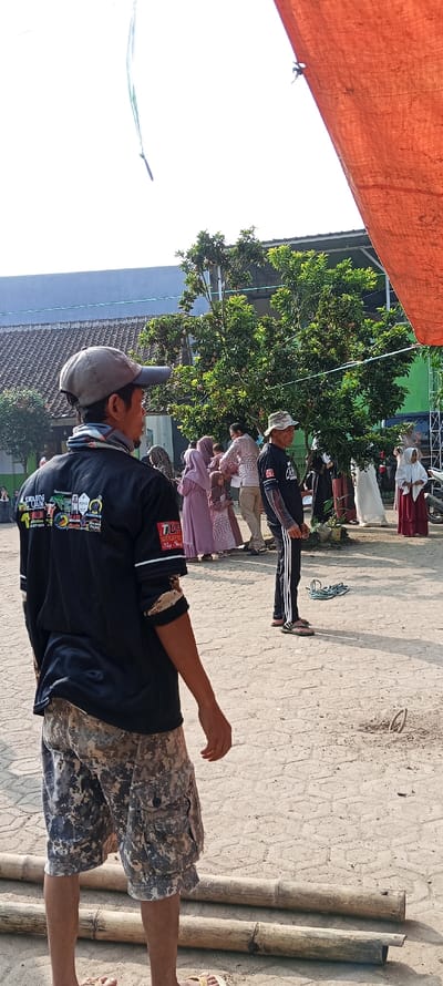 A man wearing a black shirt with numerous patches and camouflage shorts is standing in a dirt lot. He is facing away from the camera and looking to his right. Several other people are in the background of the image, including a man wearing a black shirt and white athletic pants, two women wearing hijabs, and a man and woman standing near a tree. The man in the black shirt and white pants is looking at the camera, while the other people are facing away. There are several trees and bushes in the background, as well as a red tarp hanging in the upper right corner of the frame. There are two long, thin wooden poles lying on the ground near the bottom of the image. The scene is in Cangkuang, Indonesia (latitude: -7.065, longitude: 107.552). The ground is made of gravel and dirt, and the air is clear and sunny.