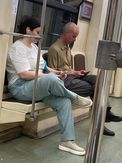 In Gavrilkovo, Russia, two individuals are seated on a train carriage. The woman on the left, wearing a white t-shirt and light blue pants, has dark hair pulled back and is using white earbuds. She is looking down, possibly at a device in her lap. Next to her, a bald man in a tan polo shirt and dark pants is also looking down at his hands, holding a small object, likely a phone. The carriage interior is visible, with beige seating and metallic poles. A digital screen displaying an orange graphic is mounted on the wall above the man. The flooring is a dark green linoleum.