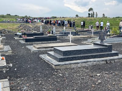 The image shows a large group of people gathered at a cemetery. They are all standing and appear to be attending a funeral. The cemetery is located on a hillside and is surrounded by trees. There are many graves, all of which are made of black granite and have a cross or star of David on top. The ground is covered with small black rocks. The mountain visible in the background is Nyiragongo Volcano. The image was taken at latitude -1.601 and longitude 29.123, which is located in Nzulo, Democratic Republic of the Congo.