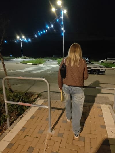A person with blonde hair, wearing a brown jacket and blue jeans, walks away from the camera on a brick path. The path leads to a parking lot illuminated by bright streetlights. A car is parked in the lot, and there is some greenery along the edge. A metal railing is visible on the left side of the path. The scene is captured at night.
