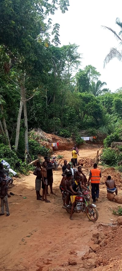 This media file captures a gathering of people, predominantly men, on a reddish-brown dirt road in a densely forested, tropical environment. The scene is set in Aloko, Nigeria, under bright daylight with a clear sky visible above the lush green canopy.
The main subjects are a group of individuals of varying ages, many shirtless or in simple attire, congregating on and around the unpaved road. Some stand casually, while others are seated, including two men on a red motorcycle with yellow detailing, and one man perched on a large rock. In the mid-ground, a man in a yellow shirt is actively gesturing or speaking to others, while further back, another man in a bright orange safety vest walks away. Two motorcycles are visible; the red one in the foreground and a less distinct one further back to the left.
The setting is characterized by thick, verdant jungle vegetation bordering the road, suggesting a rural or remote location. Mounds of reddish-brown earth flank the road, and clothes are seen drying on lines strung between trees or structures in the background, indicating human presence and daily life amidst nature. The overall atmosphere appears informal, possibly a communal gathering or a stop during travel. No visible text is present in the image.