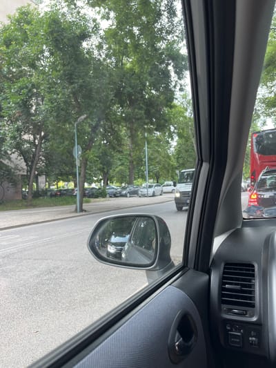 The image captures a view from inside a car, looking out the passenger side window and reflecting in the side mirror. The car is in traffic on a street lined with trees. Several other vehicles are visible, including a white van, a red bus, and various cars. The street appears to be paved and has lane markings. In the background, there are buildings and more trees. The side mirror reflects a black car and pedestrians on the sidewalk. The interior of the car is also partially visible, showing part of the dashboard and door panel. The scene is consistent with a street in the city of Orizari, Bulgaria, given the context provided.