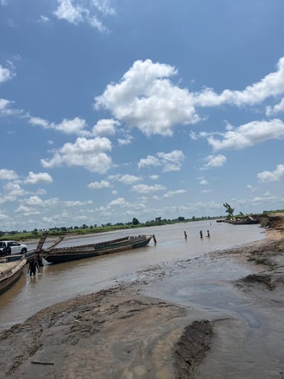 The image depicts a wide, muddy river with several wooden boats docked along the bank. The water is a murky brown, and the riverbed appears to be composed of mud and sand. In the foreground, the muddy bank is uneven with some vegetation growing out of it.
There are several boats in the water, some are partially submerged or grounded on the mud. On the left side of the image, a white vehicle, possibly a pickup truck, is parked near the boats, and two individuals are visible near the water's edge. Further into the river, in the middle ground, a group of at least three children are wading in the water. In the background, the opposite bank of the river is lined with green vegetation and scattered trees, suggesting a rural or semi-rural environment. The sky is blue with scattered white cumulus clouds.
The scene appears to be a riverbank in a natural setting, possibly a fishing or transportation hub. The presence of boats and people engaged in activities in the water indicates this is likely a functional location for the local community. The general atmosphere is bright and sunny, suggesting daytime. The specific location provided is Nemnei, Nigeria.