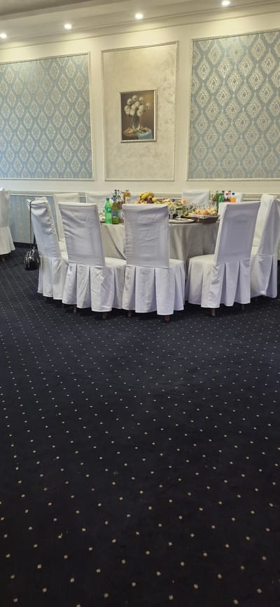 The image depicts a formal dining setting, possibly a banquet hall or restaurant, in Gavar, Armenia. The scene is oriented vertically, with chairs covered in white fabric draped over them, each featuring a ruffled skirt. These chairs are arranged around tables laden with food and drinks. On one table, a basket of fruit, including bananas and apples, is visible, alongside bottles of beverages like Sprite. Another table displays glasses and what appears to be a spread of food.
The walls are decorated with ornate paneling. The upper section features a light blue wallpaper with a repeating diamond pattern in a lighter hue. Below this, a framed painting of a bouquet of white flowers in a vase adds a touch of artistic detail. The floor is covered with a dark navy carpet dotted with small, light-colored squares.
The lighting suggests an indoor setting, possibly during the day or evening, with spotlights illuminating the upper wall sections. No people are directly visible in the frame, but the prepared tables imply an event or gathering is about to commence or has recently concluded. The overall ambiance is one of preparation for a formal occasion. No visible text is present in the image.