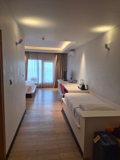 The image depicts an interior scene, potentially a hotel room, in Huraa, Maldives. The room features a bed with white linens and a wooden frame, situated on a wooden floor. Large windows are covered with curtains, allowing natural light to illuminate the space. The room also contains a sofa, and some furniture including what looks like a desk with beverages and reading materials. On a higher surface, luggage is visible. The walls are painted a light color, and lighting fixtures are installed on the ceiling. The scene appears tidy and well-maintained.