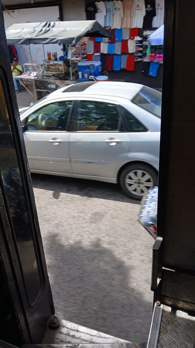 The image, taken from inside a vehicle moving on an asphalt road, shows a partially visible white car moving parallel to a small shop. The shop, located on the left side of the frame, displays a variety of colorful clothing items such as shirts, stacked in rows. Miscellaneous goods are visible behind the clothing. The shop seems to be in a commercial area in Puerto La Cruz, Venezuela, suggested by the types of merchandise and the general urban environment. Other details include parts of the capturing vehicle's interior, such as the dashboard or frame, visible at the bottom and sides of the frame. The focus is mainly on the car and the adjacent shop, and the road is partially visible on the right.