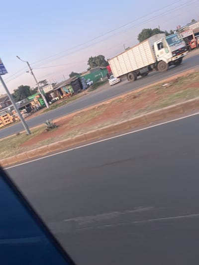 The image depicts a street scene in Biashara ward, Kenya, captured from a vehicle looking across a multi-lane paved road during what appears to be early morning or late afternoon.

**Environment:**
The foreground features a dark blue section of a vehicle, likely the camera's vantage point. An asphalt road occupies the mid-foreground, separated by a brown dirt median strip bordered by a low concrete curb. A single agave plant grows within this median. A utility pole with a streetlamp and overhead wires stands on the left side of the road. The background reveals a commercial strip with various structures and sparse vegetation under a clear, pale blue sky with slight warm hues near the horizon.

**Subjects and Objects:**
On the paved road, a large white box truck with a prominent ribbed cargo container is in motion, heading from right to left. A smaller white passenger sedan follows behind the truck. Along the roadside, a series of mixed commercial buildings and makeshift structures are visible. These include a multi-story building with exposed blockwork on the far left, followed by single-story shops or shacks constructed from various materials, including corrugated metal and green tarpaulin. One green structure clearly displays "Safaricom" branding, and another sign for "Heineken" is visible.

**People and Actions:**
Several individuals are present near the commercial establishments. Some are seated at outdoor tables and chairs, while others appear to be standing or walking in the vicinity of the shops. Specific interactions are indistinct due to distance and image resolution, but the scene suggests routine daily activity.