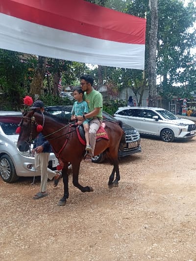The scene is set in Kajar, Indonesia, depicting a brown horse with a red saddle and a red pompom on its head, moving rightward across a light brown gravel surface. An adult male, wearing a green t-shirt and glasses, is seated on the horse behind a child in a patterned blue shirt. The adult's right arm is around the child. A male handler, dressed in a dark blue shirt and light-colored pants, walks on the horse's left side, holding the reins. A large red and white Indonesian flag is prominently displayed horizontally across the upper portion of the frame. In the background, three vehicles are parked: a silver hatchback on the far left, a dark SUV directly behind the horse, and a white SUV further to the right. The environment is characterized by dense green foliage, numerous tree trunks, and indistinct structures in the distant background. Several additional individuals are faintly visible in the mid-ground and background.