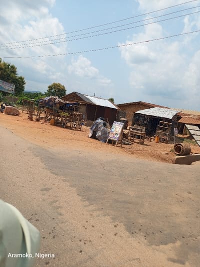 The image shows a dusty road with a few small buildings on the side. There are three structures to the right of the road, one of which is a partially obscured small shop. The shop has signs in front of it, and the image suggests that it is open for business. The road is surrounded by dirt and dirt piles, and there is a slight bend in the road near the camera. The location of the image is Aramoko, Nigeria, based on the coordinates provided. The sky is cloudy and there are power lines over the road. The image is taken from the point of view of someone looking down at the road.