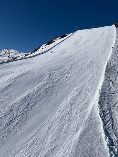 The image depicts a wide, snow-covered ski slope under a clear, deep blue sky, located in the city of Schwendau, Austria. The slope, primarily white with extensive ski tracks, is illuminated by direct sunlight. In the mid-ground, a single individual wearing red ski attire is skiing downhill towards the lower left of the frame. Further up the slope, toward the right, several small figures, likely skiers, are visible near a ski lift line. A ski lift structure, including chairs, is partially visible at the top right edge of the image. Along the upper left portion of the slope, a red safety netting or barrier runs horizontally below a darker, rocky mountain section. Distant snow-capped peaks and additional ski lift towers are discernible in the far left background. Yellow marker poles delineate a specific section of the groomed slope on the right side. The scene is devoid of explicit interactions between individuals.