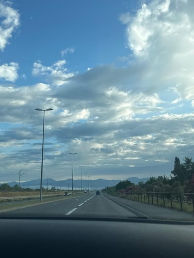 The image depicts a daytime scene captured from inside a moving vehicle on a multi-lane highway. The road, with clear white lane markings, recedes into the distance, where several cars are visible, including a dark-colored vehicle in the mid-ground.
To the left of the highway, a fence or barrier separates the road from a dense line of trees and bushes, some displaying reddish foliage, hinting at autumn or specific plant types. Beyond this greenery, a range of hazy mountains stretches across the horizon, forming the backdrop of the scene.
On the right side of the highway, a series of tall streetlights with dual fixtures extends along the road, diminishing in size towards the vanishing point. Utility poles and power lines are also present in this area.
The upper half of the image is filled with a dynamic sky. A vibrant blue sky peeks through numerous white and grey clouds, which vary from fluffy cumulus shapes to elongated stratocumulus formations. The clouds are well-lit, suggesting either morning or late afternoon sunlight.
The setting appears to be a picturesque highway journey through a mountainous or hilly region, consistent with the Goričani, Montenegro location. No specific human activity beyond driving is evident, and no visible text is present.