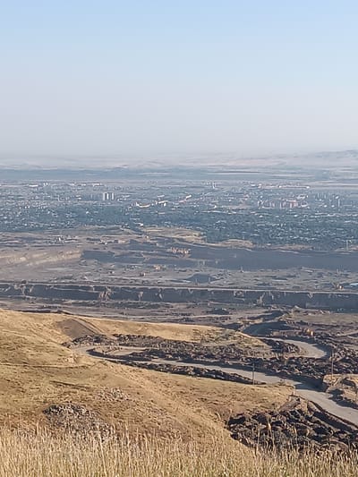 The image shows a wide, panoramic view of a city in the distance, possibly Almalyk, Uzbekistan, under a clear, hazy sky. In the foreground, a dry, grassy hillside slopes down towards a large, excavated area. This excavated area appears to be a mining operation, with terraced earth and dark soil layers visible. Several dirt roads wind through this industrial zone. A few scattered pieces of heavy machinery, small yellow construction vehicles, are visible within the excavation. The city in the background is densely populated with numerous buildings of varying heights, interspersed with trees and greenery. The overall scene suggests an industrial landscape adjacent to a residential or urban area.