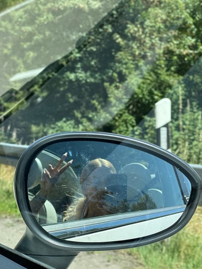 The image is a reflection in a car's side mirror, showing the driver taking a selfie while holding up a peace sign. The background visible through the car's windows and in the mirror is a wooded area with greenery. The driver has blonde hair and is wearing a light-colored top. The car appears to be in motion, as suggested by the blurred scenery. The location is the city of Furtwangen im Schwarzwald, Germany.
