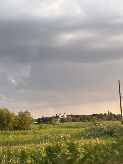 The image depicts a rural landscape in Soglasny, Russia, with a dramatic sky dominating the left side. Heavy, dark gray clouds suggest impending or ongoing bad weather, possibly a thunderstorm, with lighter patches indicating breaks in the cloud cover. A telephone pole with wires stretches across the upper portion of the frame, signifying the presence of civilization in this otherwise natural setting.

To the right, a lush green field stretches towards a line of trees that forms the horizon. The sunlight, though partially obscured by clouds, illuminates the field, creating stripes of light and shadow, and highlighting the texture of the tall grass. In the middle ground, nestled among the trees, a white building with a dark dome, likely a church or monastery, is visible. This religious structure adds a cultural and historical element to the scene.

The overall mood is one of atmospheric intensity and natural beauty. The contrast between the dark, foreboding sky and the vibrant, sunlit field creates a dynamic visual tension. The time of day is likely late afternoon or early evening, given the low angle of the light and the dramatic sky. The location cues of a rural field, a church, and power lines are consistent with a setting in Soglasny, Russia. No text is visible in the image.