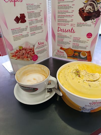 The image showcases a cafe setting with a menu and food items. The menu is open, displaying various desserts and crepes. Prominently featured on the left is a strawberry crepe with chocolate sauce and a scoop of pink ice cream, labeled as "Strawberry Dream." To the right of the menu is a selection of desserts including a chocolate cake with ice cream, cheesecakes, and cookie doughs.
In the foreground, a white cup of coffee with latte art sits on a saucer. Beside the coffee cup is a small white packet, likely containing sugar or creamer. Partially visible in the lower right corner is a large tub of yellow ice cream with chopped nuts, branded as "Anjoma Persian Saffron Ice cream."
The scene appears to be indoors, likely a cafe or dessert shop. The lighting suggests it could be daytime. The overall mood is relaxed and inviting, highlighting the sweet treats available. Visible text includes item names and prices on the menu, as well as branding on the ice cream tub.