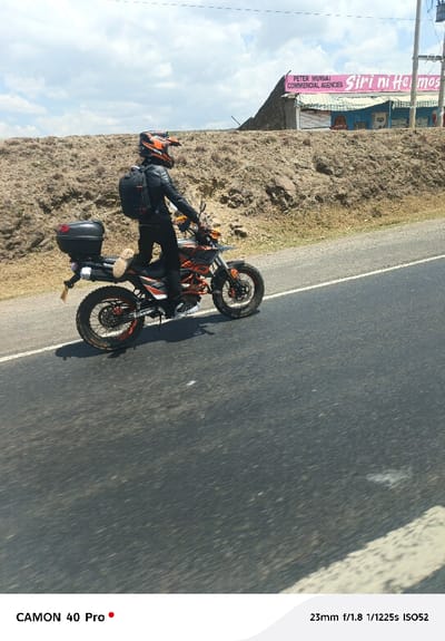 The image depicts a single individual on a motorcycle positioned on the paved shoulder of a road in Gilgil ward, Kenya. The person is dressed in a black leather jacket, black pants, and an orange, black, and white full-face off-road helmet. A black backpack is worn on their back. The motorcycle is an orange, black, and white enduro/dual-sport style, equipped with knobby tires, orange wheel rims, and a black hard-shell top case mounted on the rear rack. One foot, wearing a light-colored shoe, is visible resting on the motorcycle.

The road surface is dark asphalt with a white solid line delineating the shoulder. Immediately behind the road is a steep, reddish-brown dirt embankment. On top of this embankment, in the background, two commercial buildings are visible. The building on the left features a pink upper facade with white text reading "PETER MUKAI COMMERCIAL AGENCIES." The adjacent building to its right is light blue with "Siri ni Hemo" partially visible on its sign. Utility poles and wires are present in the far background. The sky above is partly cloudy with patches of blue visible.