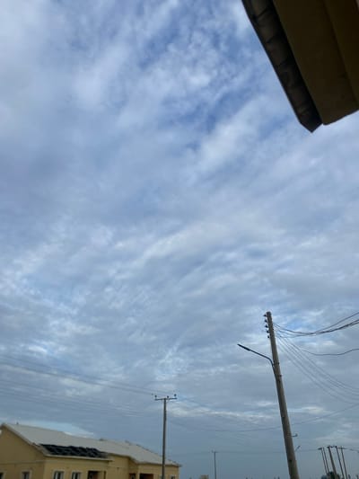 The image depicts an outdoor scene under a cloudy sky. In the lower left, a portion of a pale yellow, single-story building with a white roof is visible. The building appears to be under construction or renovation, with a section of the roof showing dark, open beams. Several utility poles with power lines crisscross the sky above the building. One prominent wooden utility pole stands in the mid-right of the frame, carrying numerous wires and a street lamp. Another utility pole with a street lamp is visible further in the background on the right. The sky is filled with a textured layer of grey and white clouds, suggesting an overcast or possibly approaching rainy weather. The scene appears to be in a semi-urban or developing area, consistent with a location in Dumnga, Nigeria. No people or animals are present in the image.