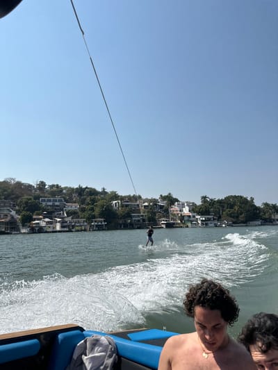 A water skier is towed by a boat across the water near a shoreline with houses. The sky is bright blue. Two people are on the boat, a curly-haired man in the foreground and another person behind him. The boat's blue interior is partially visible. The water skier creates a wake as they move.