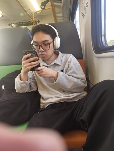 A young East Asian male, wearing glasses and white headphones, sits on a train, looking at his smartphone. He is dressed in a light gray sweatshirt and black pants. Next to him is a black backpack. The train interior is visible with seats, windows, and overhead lighting. The image appears to have been taken on a sunny day. The location is not identifiable from the image.