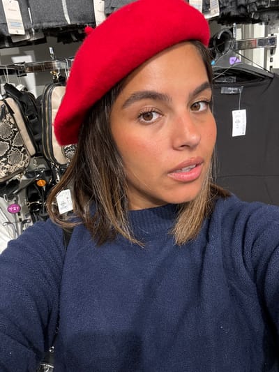 A young adult female is depicted in a close-up selfie within a Primark retail store, located at 1600 Market Street, Philadelphia, United States. She wears a red beret and a navy blue knitted sweater. Her brown hair, featuring lighter streaks, frames her face. Her eyes are brown, and her expression is neutral. The background details include various retail fixtures and merchandise. To the left, hanging handbags are visible, one prominently displaying a snakeskin pattern. Above these, gray textile items, possibly scarves, with "Primark" and "$8" price tags are present. To the right, a black garment on a hanger bears a "Primark" tag indicating size "XL" and a price of "$17.00". The environment is an illuminated indoor commercial space.
