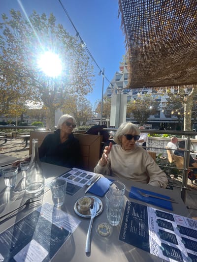 The scene depicts an outdoor dining area in La Grande-Motte, France, under a clear, bright sky. Two elderly women are seated at a rectangular grey table in the foreground. The woman on the left has short white hair and wears dark sunglasses and a dark long-sleeved shirt. The woman on the right has short white hair, wears dark sunglasses, and a light-colored long-sleeved top; she is actively gesturing with both hands, suggesting conversation.
On the table are multiple clear drinking glasses, an empty glass carafe, several blue and white menus written in French, blue folded napkins, silverware (forks and knives), and small dishes containing food items—one with a light-colored spread and a small fork, another with a ring-shaped item. Intense sunlight creates a bright glare on the left side of the table.
The environment includes trees with sparse, yellowish leaves, suggesting late autumn or early spring. Overhead, a light-brown mesh shading material is partially visible, strung with decorative lights. In the background, distinctive white, pyramidal architectural structures, characteristic of La Grande-Motte, are prominent. Other patrons and a person in a white shirt (possibly staff) are vaguely visible in the background, seated at additional outdoor tables.
